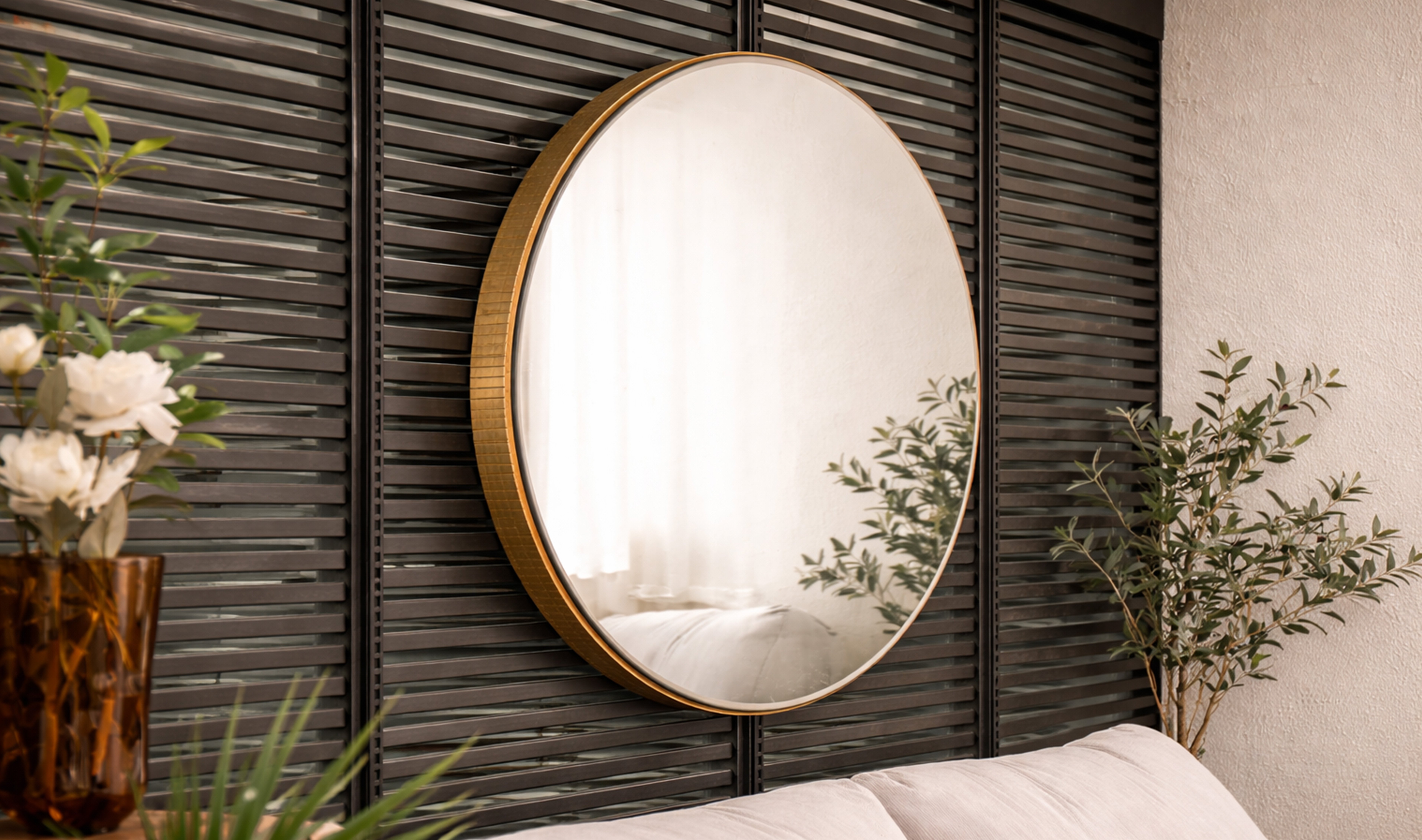 Round gold-framed wall mirror above a light sofa, against dark slatted panels with green plants.